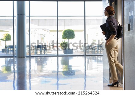Businesswoman exiting elevator into lobby, carrying shoulder bag and folder, side view