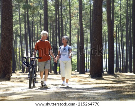 Senior couple in cycling helmets walking through wood with bicycles, holding hands, front view