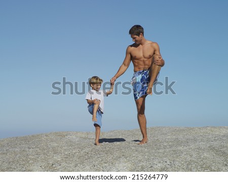 Father and son (4-6) playing on rock, standing on one leg, smiling, man in swimming shorts