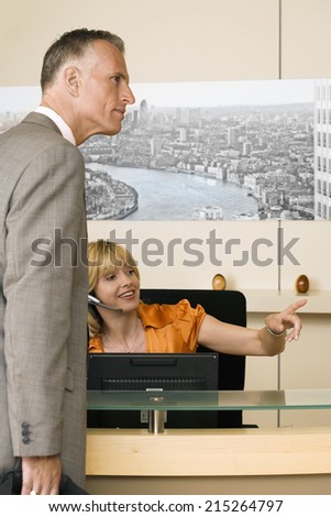 Receptionist working behind reception desk, showing businessman the way, pointing finger, smiling