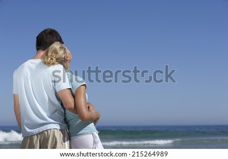 Couple standing on beach, looking at horizon over sea, man embracing woman, rear view