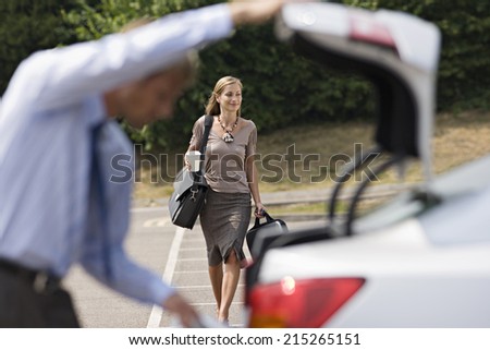 Man opening car boot in car park, focus on businesswoman walking with luggage in background, smiling