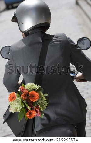 Man in crash helmet riding on scooter in street, carrying flower bouquet in bag, rear view (tilt)