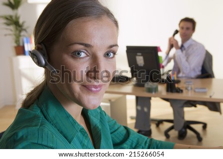 Man at desk in office, using telephone, focus on businesswoman with hands-free device in foreground