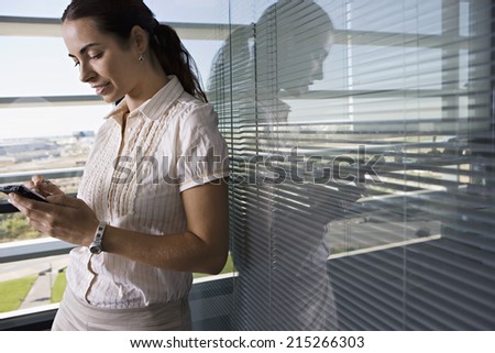 Businesswoman leaning against office window, using personal electronic organiser, side view