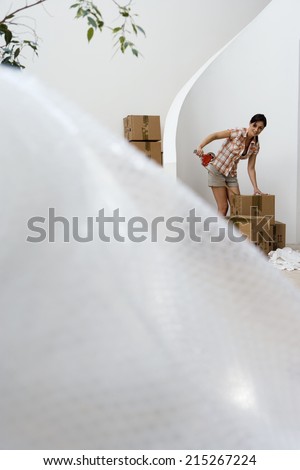 Woman sealing box with tape at home, smiling, bubble wrap in foreground, focus on background
