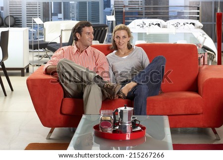 Couple sitting on new red sofa in furniture store, man glancing at woman, smiling, portrait