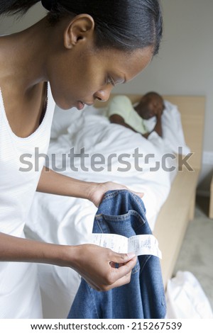 Man sleeping in bed, focus on woman holding pair of jeans and receipt in foreground, side view