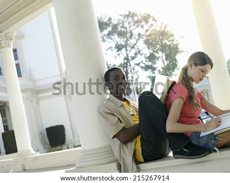 University students studying near colonnade, man leaning book against woman\'s back, smiling (tilt)