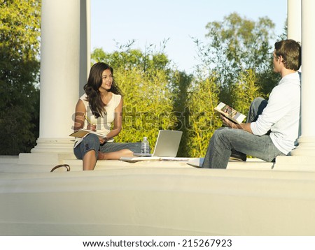 Two university students studying near colonnade, woman beside laptop and textbooks, smiling