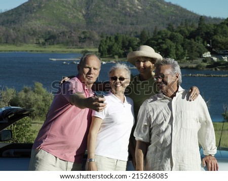 Two senior couples standing beside car near lake, man taking photograph with digital camera, smiling