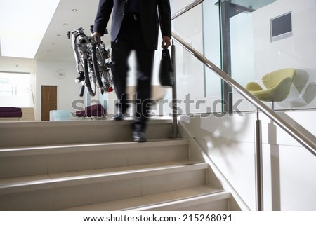 Businessman carrying commuter bicycle up steps in lobby, low section, rear view (blurred motion)