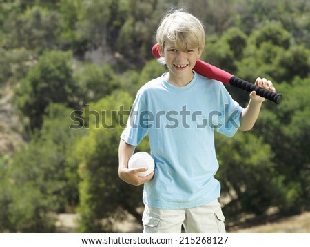 Blonde boy standing in park with softball bat and ball, smiling, front view, portrait