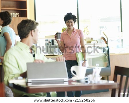 Man with laptop sitting at cafe table, greeting young woman carrying mug of coffee, smiling