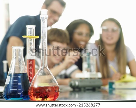 Teenagers (15-17) doing science experiment, teacher assisting, focus on conical flasks in foreground