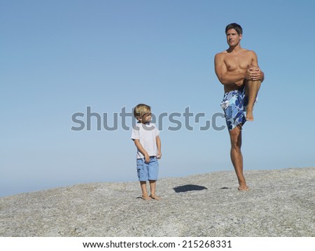 Father and son (4-6) playing on rock near beach, smiling, man in swimming shorts standing on one leg