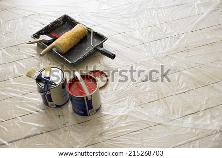 Paintbrushes, tins of paint, roller and tray on plastic sheet on wooden floor (still life)