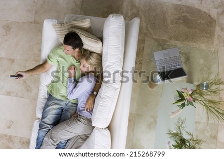 Couple relaxing with feet up on sofa at home, watching tv, man holding remote, overhead view