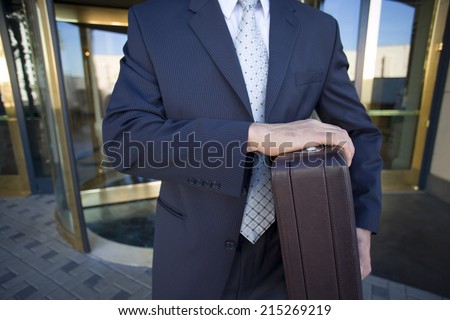 Businessman standing in front of revolving door, carrying briefcase, front view, mid-section
