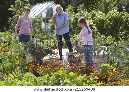 Girl (8-10) watering flowers in garden, mother and grandmother looking on, smiling, side view
