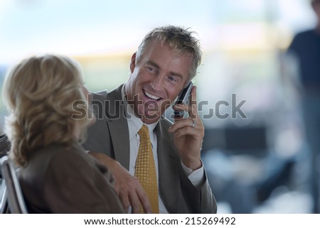 Businesswoman and businessman sitting in waiting area, man using mobile phone, smiling, side view