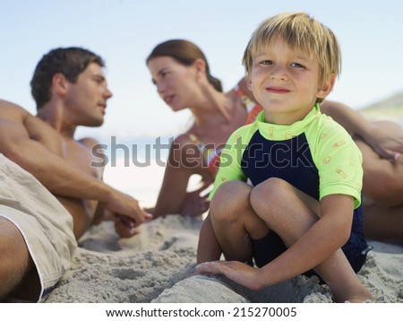 Family sitting on beach, focus on boy (5-7) crouching in foreground, smiling, portrait