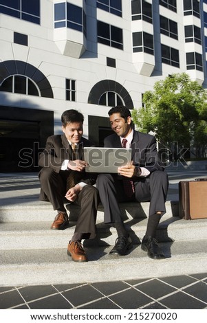 Two businessmen using laptop on pavement steps outside office block, smiling