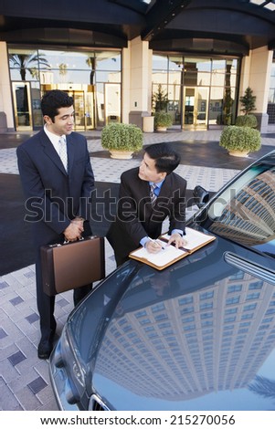 Two businessmen standing beside car outside hotel, man signing contract, smiling