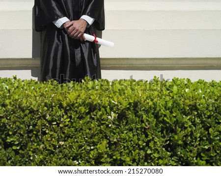 University student in graduation gown holding diploma, mid-section, hedge in foreground