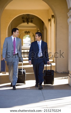 Two businessmen walking side by side in building arcade, luggage in tow, talking, front view