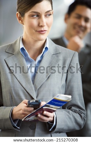 Businesswoman standing in airport terminal, holding mobile phone and ticket, making sideways glance