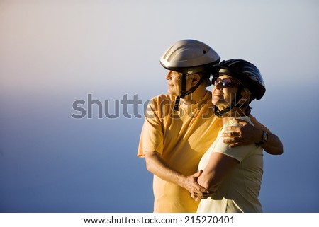 Senior couple, in cycling helmets, embracing on clifftop, looking at Atlantic Ocean horizon, side view