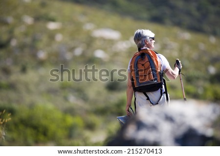 Mature woman hiking on mountain trail, carrying rucksack and using hiking pole, rear view