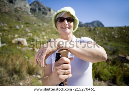Mature woman, in hat and sunglasses, hiking on mountain trail, leaning on hiking pole, smiling, portrait