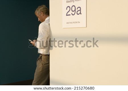 Businessman leaning against wall outside meeting room, making private call on mobile phone, side view