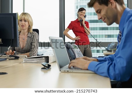 Three business people working in office, man using laptop, woman using PC, second woman using phone