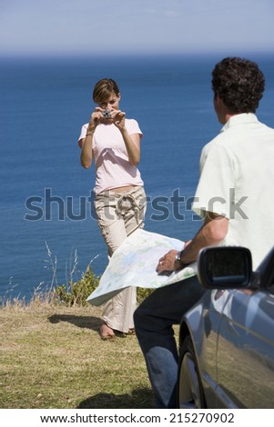 Couple standing on clifftop overlooking Atlantic Ocean, woman photographing man with map beside car