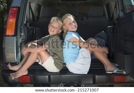 Boy and girl sitting in car boot, back to back, smiling, side view, portrait