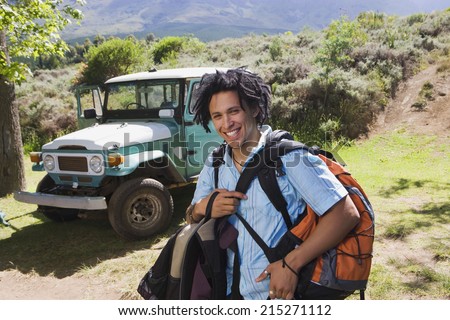 Young man unloading parked jeep at start of camping holiday, carrying rucksacks, smiling, portrait