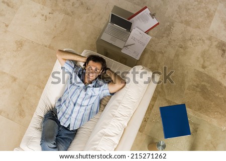 Man lying on sofa beside laptop on coffee table at home, hands behind head, smiling, overhead view