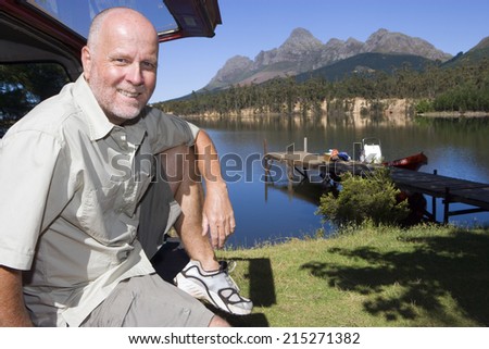 Mature man sitting in boot of parked SUV, smiling, side view, portrait, lake and jetty in background