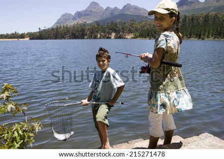 Brother and sister (7-10) fishing in lake, boy holding fish in net, girl holding rod, smiling, portrait