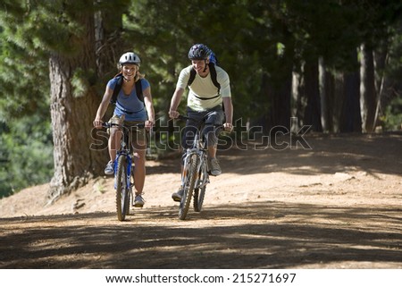 Couple, with rucksacks and cycling helmets, mountain biking along woodland trail, smiling, front view