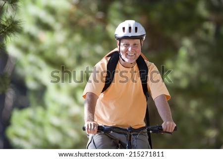 Senior man, in orange t-shirt and cycling helmet, mountain biking on woodland trail, smiling, front view, portrait