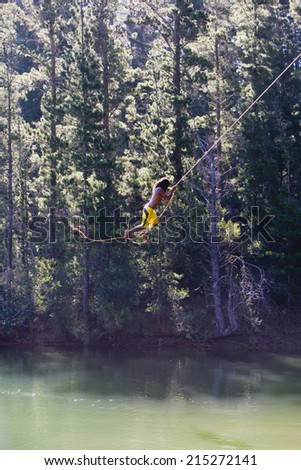 Man, in yellow swimming shorts, swinging on rope above lake, side view