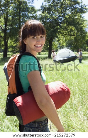 Young woman carrying rucksack and sleeping bag to camp in woodland clearing, smiling, portrait, friends assembling tent