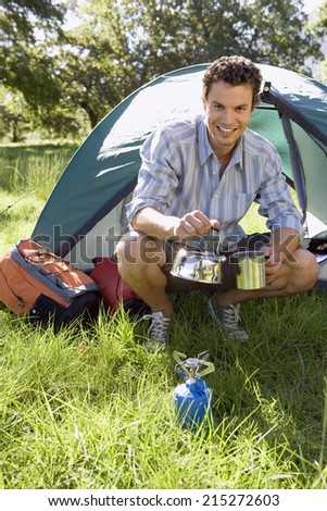 Young man crouching beside dome tent in woodland clearing, taking boiled kettle from camping stove, smiling, portrait