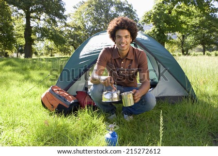 Young man crouching beside dome tent in woodland clearing, taking boiled kettle from camping stove, smiling, portrait