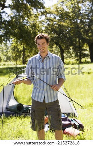 Young man assembling dome tent on camping trip in woodland clearing, smiling, portrait