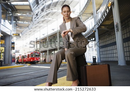 Businesswoman sitting on suitcase, waiting for city tram, checking time on wristwatch (surface level)